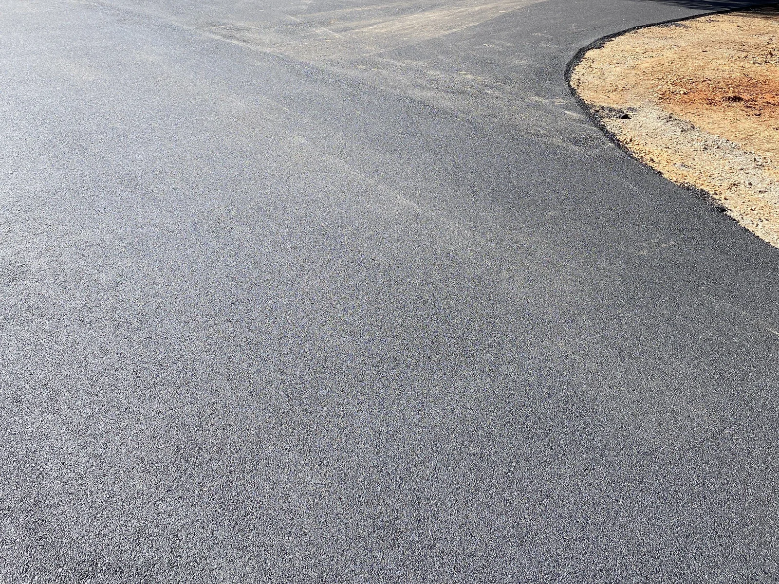 Freshly rolled asphalt driveway curving past a gravel shoulder toward a fenced yard