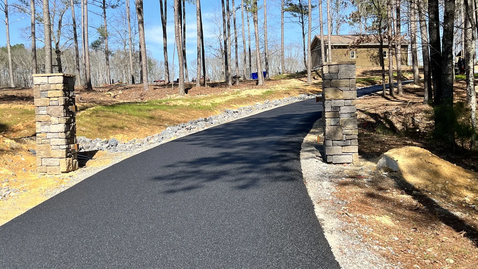 Fresh asphalt driveway passing between stone entry columns into a stand of pines
