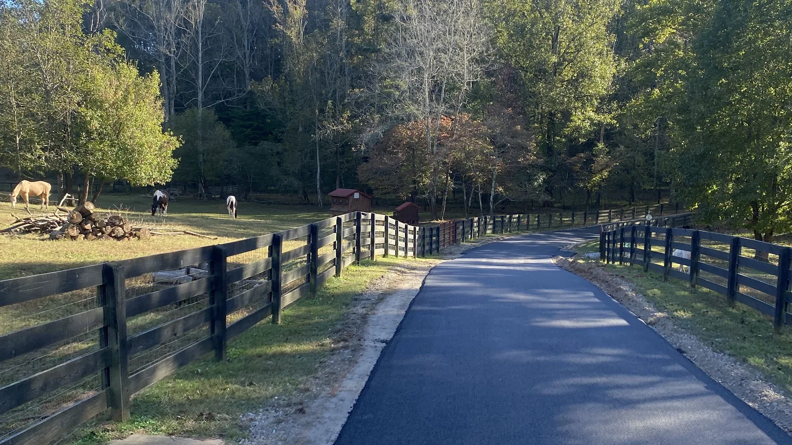 Asphalt paving in Ball Ground, GA: a new asphalt lane between black board fences past a horse pasture toward the tree line