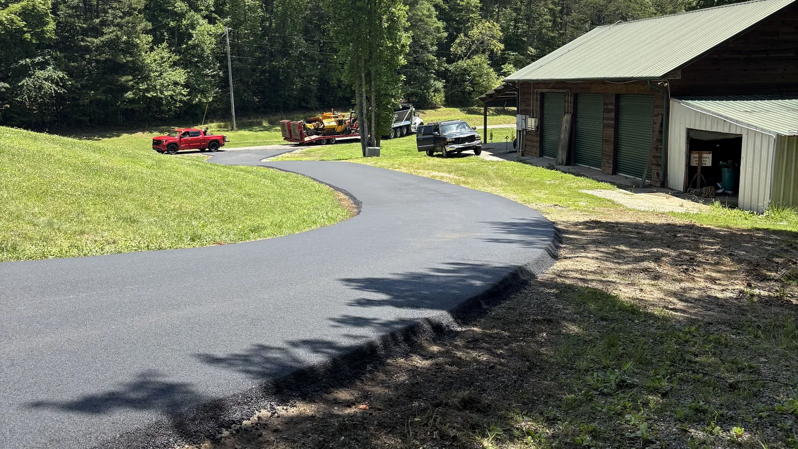 Asphalt paving in Blairsville, GA: a new asphalt driveway curving across a lawn toward a metal-roofed barn with tall pines behind it