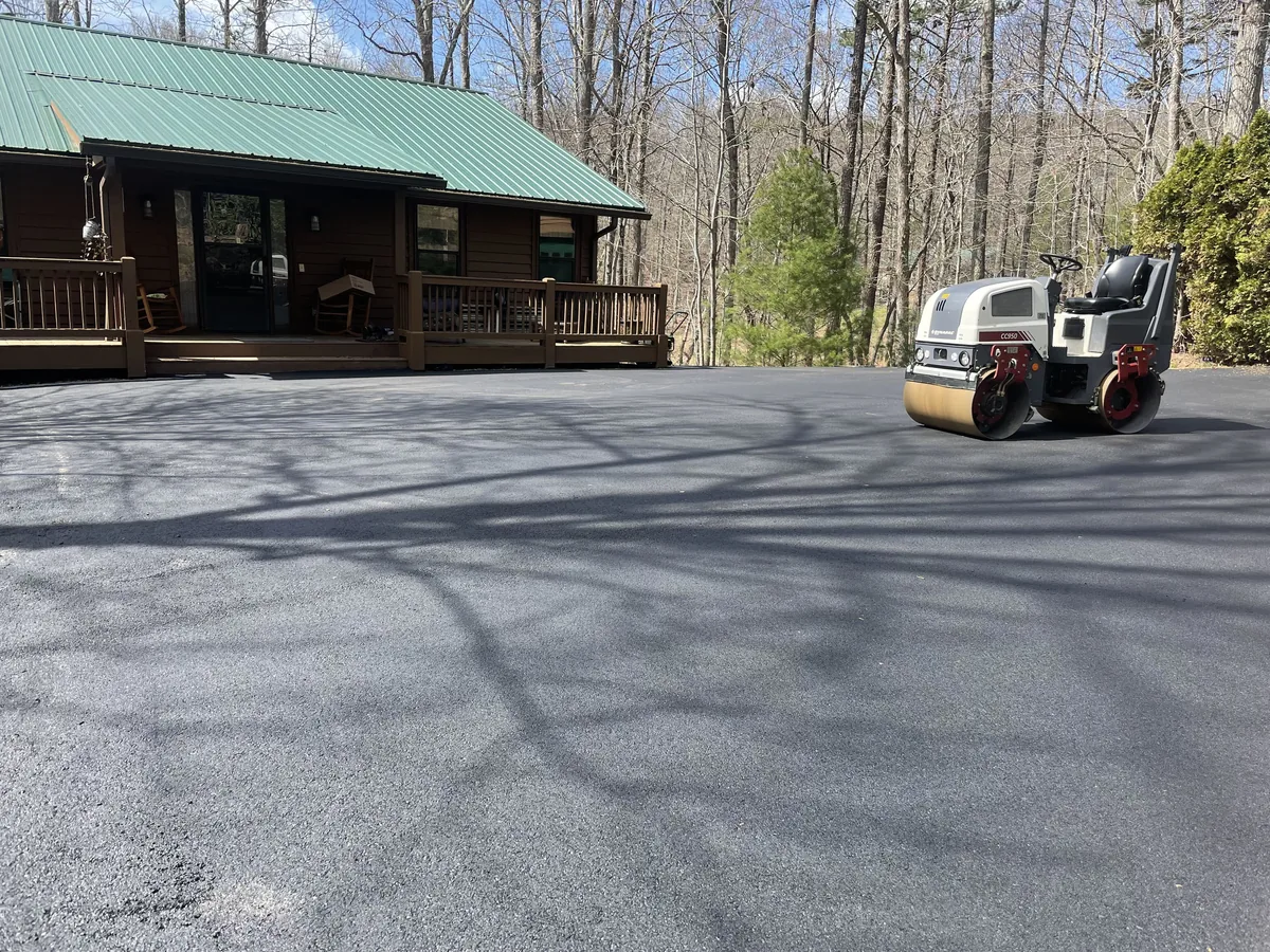 Ride-on roller compacting a freshly laid asphalt driveway beside a cabin