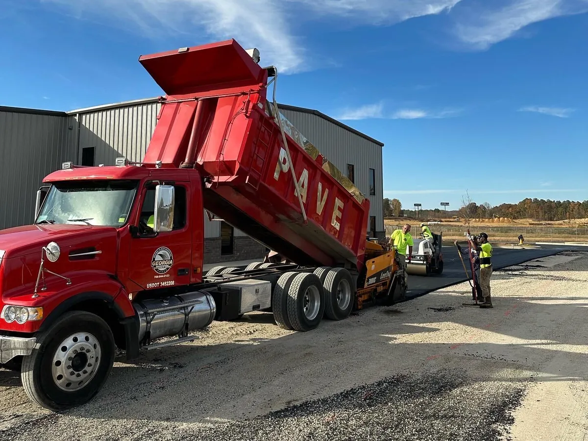 Red dump truck with its bed raised feeding an asphalt paver while the crew works behind it