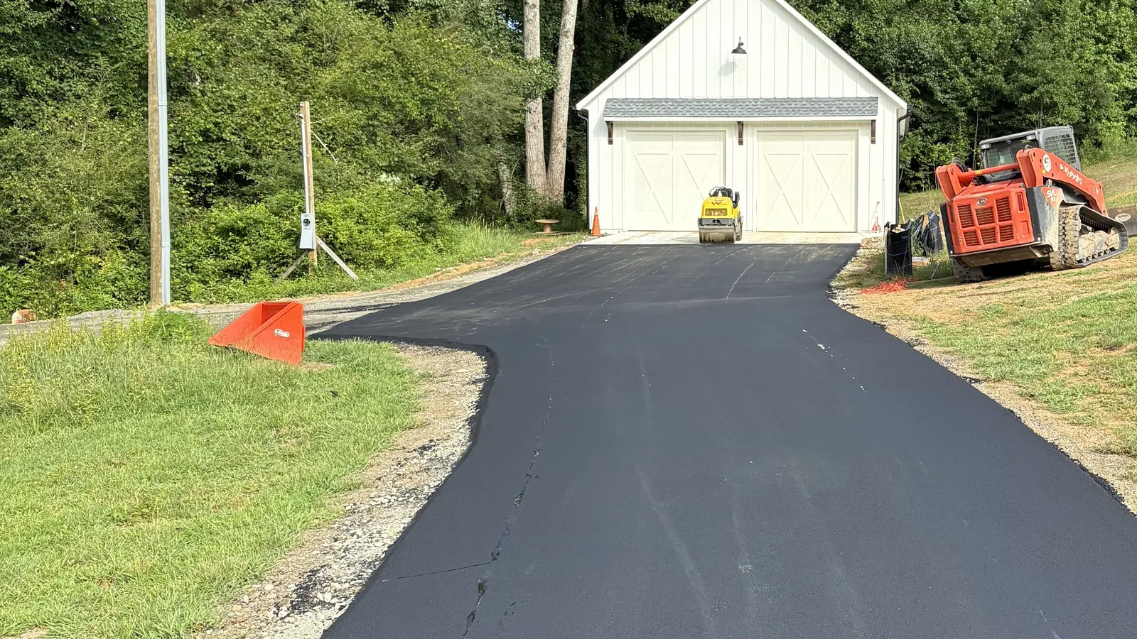 Asphalt paving in Jasper, GA: a fresh asphalt driveway running up an open lot to a white two-bay garage, the track loader and roller at the edge