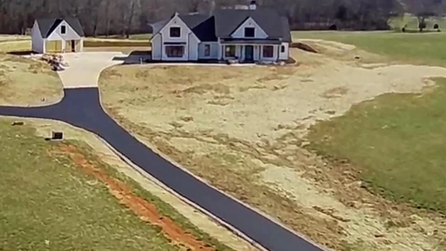 Aerial view of a long new asphalt drive crossing a pasture to a white farmhouse