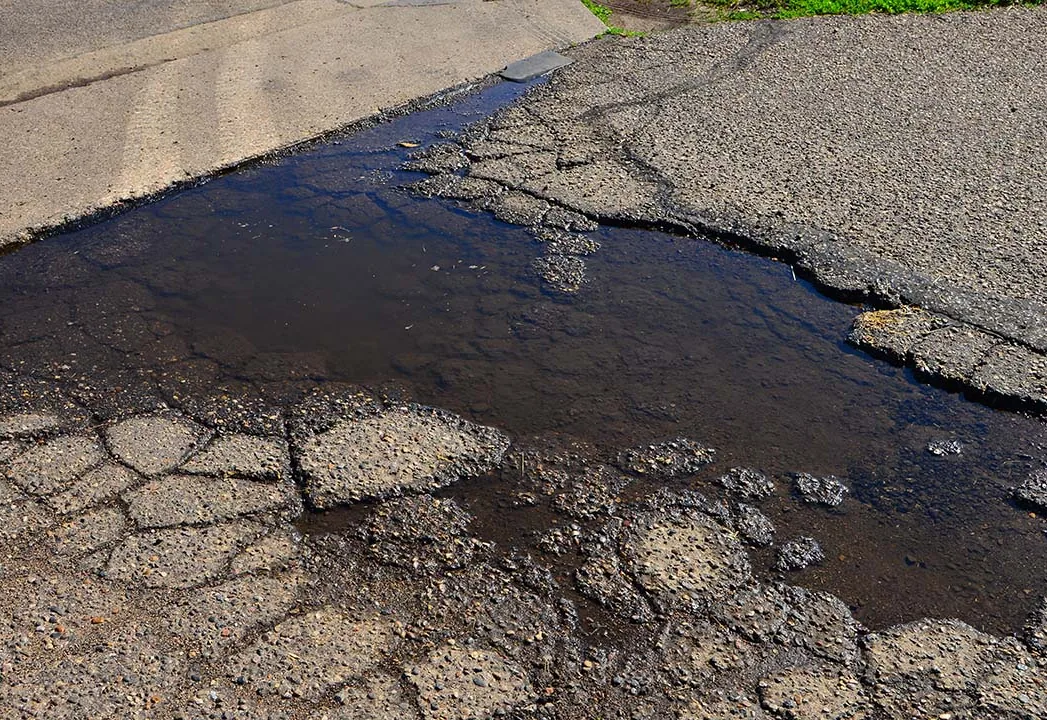 Broken, crumbling asphalt with standing water pooling in a pothole at a concrete gutter