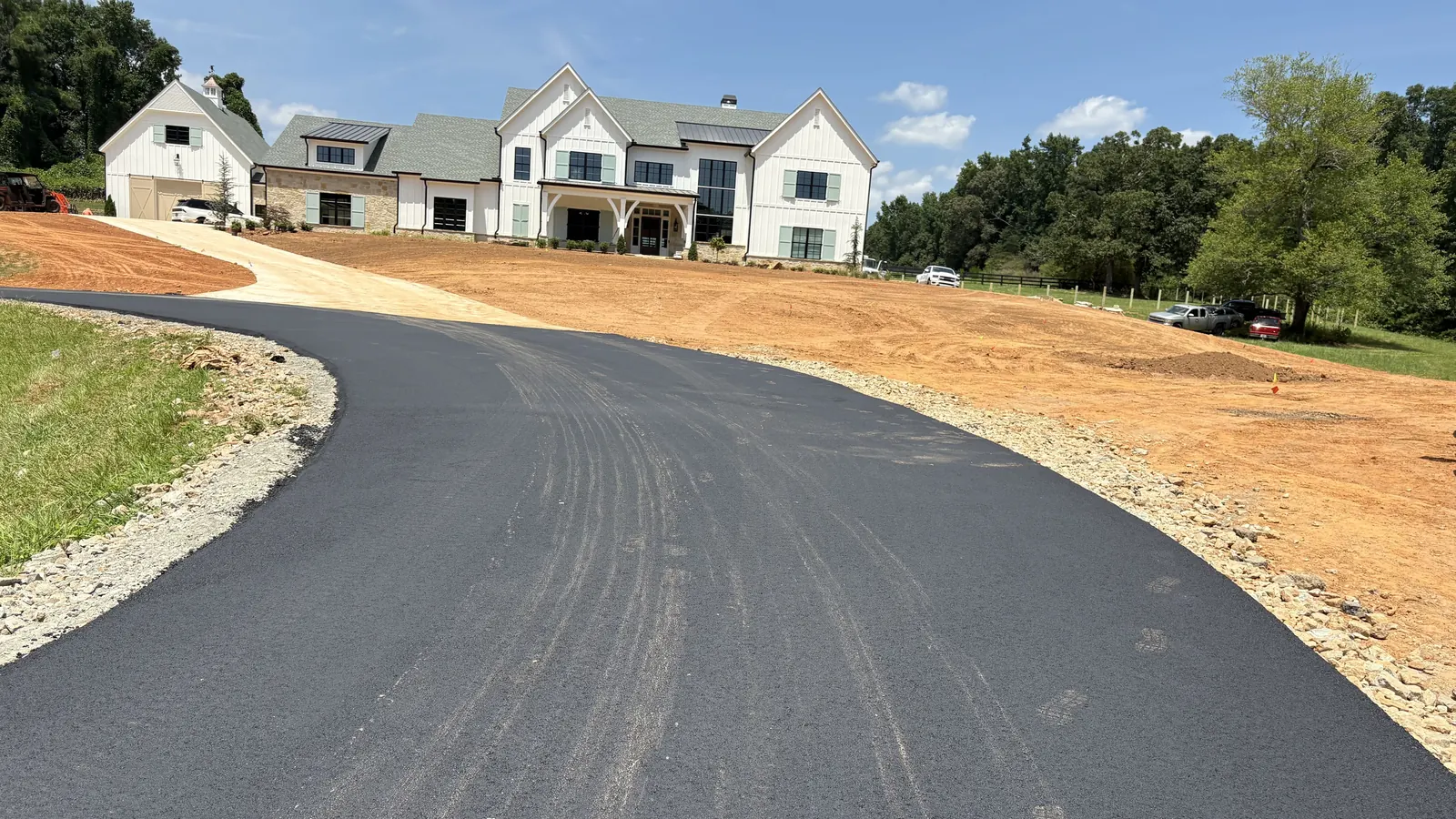 Wide curving driveway of fresh black asphalt leading to a white farmhouse across a green pasture
