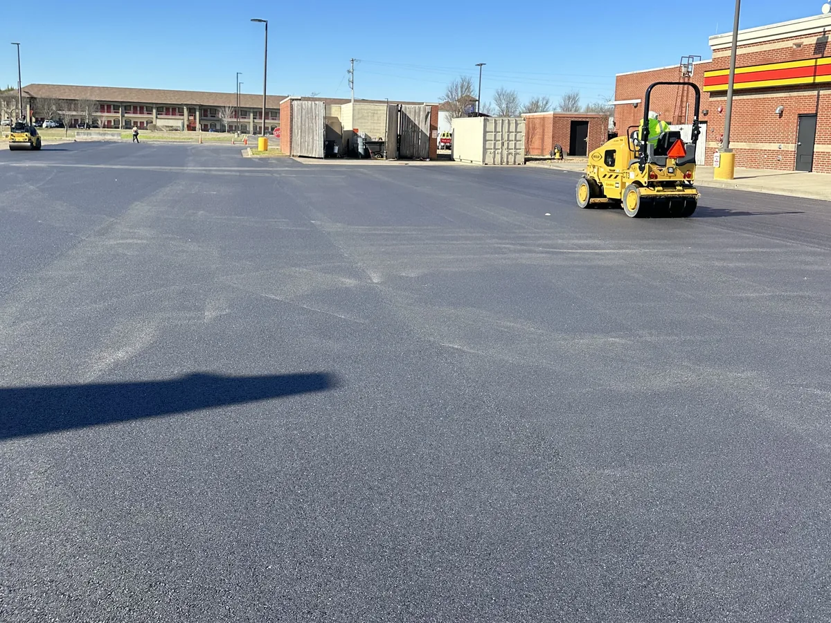 Roller compacting fresh asphalt on a commercial parking lot behind a row of buildings