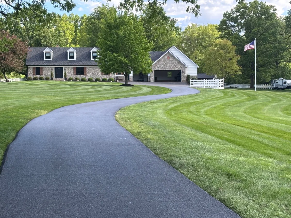 Freshly sealcoated black driveway curving across a striped green lawn to a house
