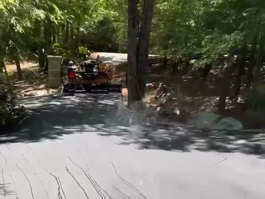 Sealer being spray-applied in overlapping fans across an older asphalt driveway in the woods